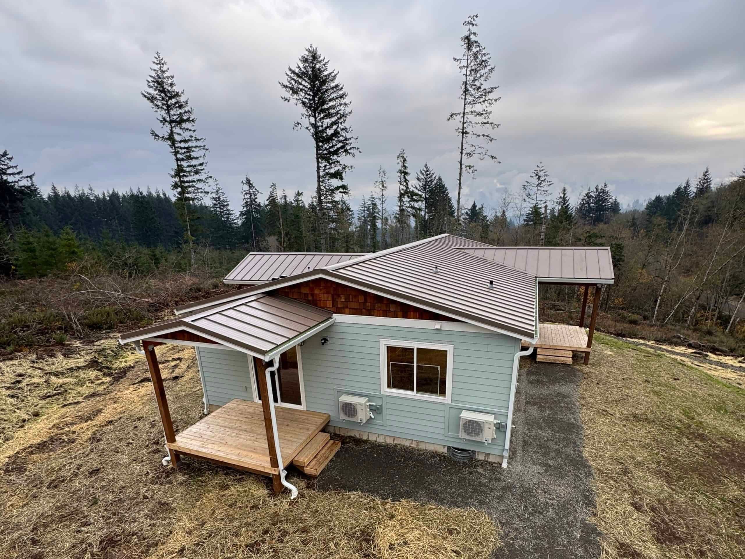 Light-blue Wolf Model I with covered wood porches and a standing-seam metal roof on a wooded hillside in Battle Ground