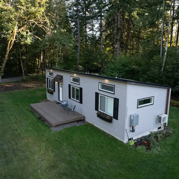 Finished light-gray Wolf home with black window trim and a wood deck on a lawn among tall trees