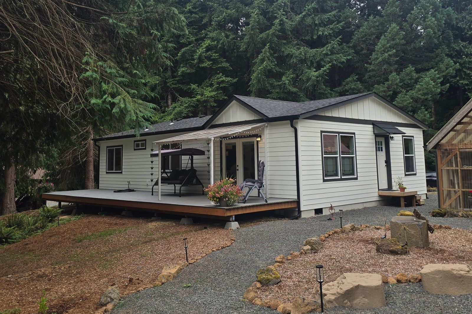 White Wolf Model I with black trim, a furnished deck, and a landscaped gravel path among evergreens