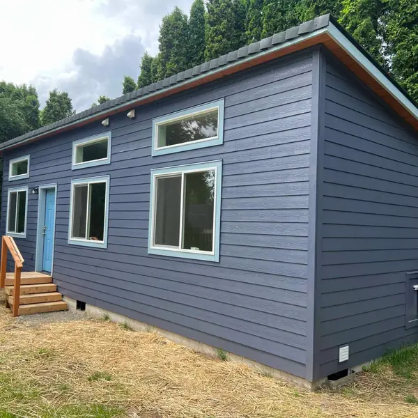 Slate-blue Wolf Model K with pale-blue window trim, a light-blue door, and a wall-mounted heat pump among evergreens