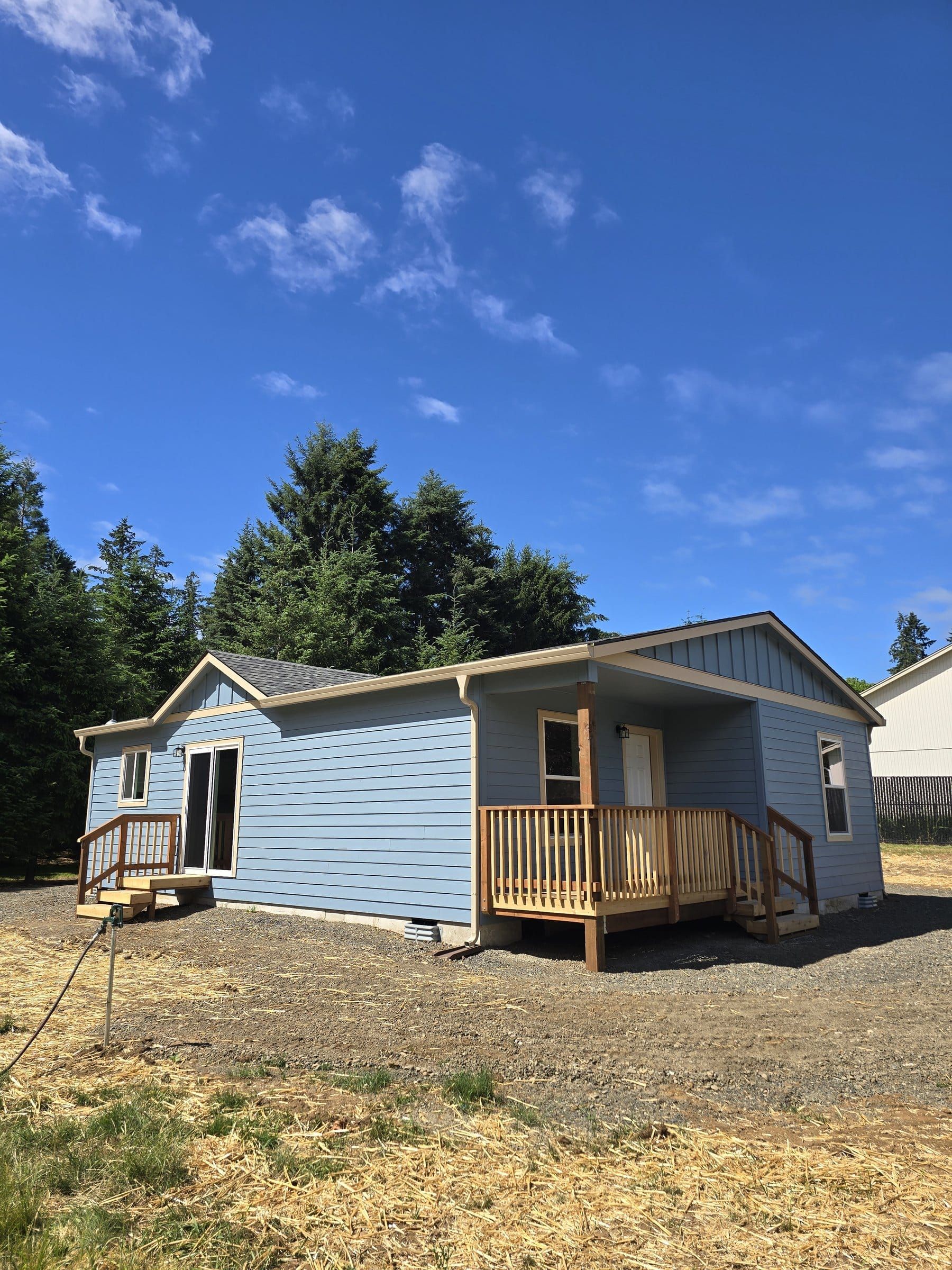 Light-blue Wolf Model L with a covered wood porch, a sliding-door deck, lap and board-and-batten siding, and evergreens behind it